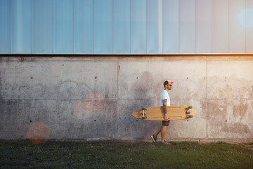 Hipster in white blank t-shirt, shorts, sneakers, baseball cap and sunglasses carrying a longboard against an industrial city building background with lens flare