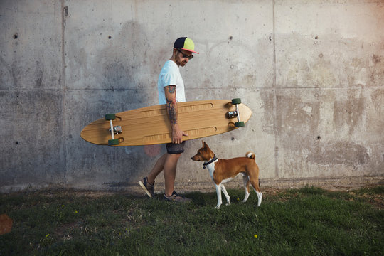 Young surfer in unlabeled white t-shirt looking down at his dog as he walks along a gray concrete wall