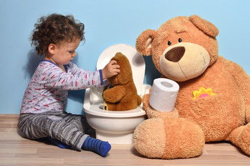Baby toddler sitting on the floor teaching a small teddy bear who sits on a potty. Cute kid potty training for pee and poo helped by teddy bear who gives him toilet paper - obrazy, fototapety, plakaty