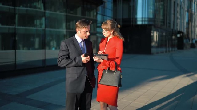 Two young attractive businesspeople. Businessman in suit and tie watch in smart phone. Businesswoman try to make offer with digital pad but he reject her offer. Glossy Business centre district bg. 4k