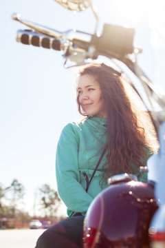 Portrait of young attractive woman with long curly hair wearing green sweater and jeans is sitting on red motorcycle and looking aside on a road on summer day outdoor. Lifestyle, vacation, joy concept