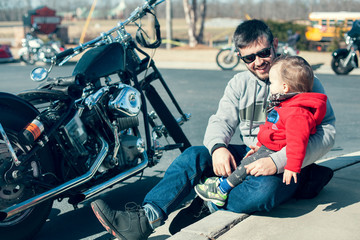 Handsome young father and cute little toddler son sitting on the ground and looking on each other near black motorcycle on summer day outdoor. Lifestyle, vacation, happiness, joy concept