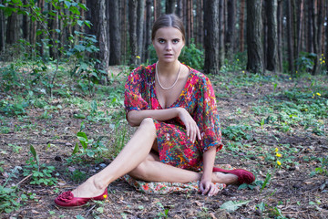 Young beautiful model in a red dress is sitting on the ground in the forest