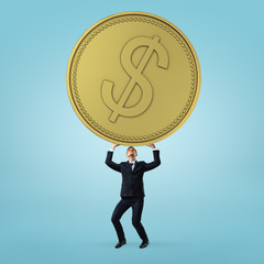 Businessman stands and holding heavy, big golden coin on blue background