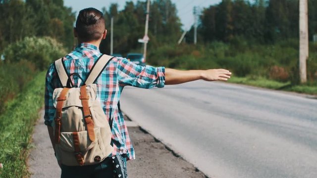 Back side young boy with backpack hitchhiking at road in summer sunny day. Thumb. Cars. Countryside.