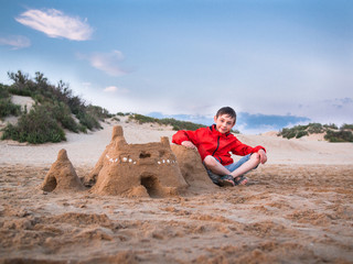 Little boy in shorts and a jacket sitting legs crossed near the sandy castle against the blue sky and the sand dunes with bushes