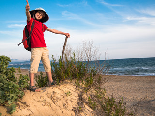 Toned image of a little boy with a backpack and a hat that holds the stick and standing on a sandy hill and smiling against the background of the calm sea and clear sky