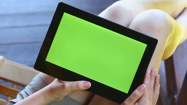 Young woman sitting on wooden chair browsing internet and watching video on touch pad. Using tablet pc with blank green screen. Point of view video footage of hands of anonymous woman and touchpad.