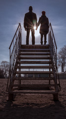 Toned image of man and woman holding hands and standing at the top of a rusty iron ladder against the clear sky and the sun backlight