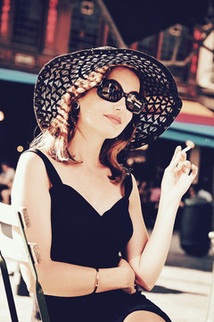 jolie femme brune style années 1960 en robe noire et chapeau vintage fumant sur une terrasse au soleil