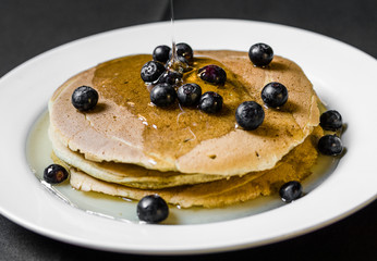Close up of a bunch of tasty american pancakes with colorful blueberries and maple syrup poured over them. Selective focus.