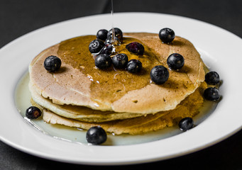 Close up of a bunch of tasty american pancakes with colorful blueberries and maple syrup poured over them. Selective focus.