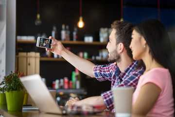Happy romantic couple posing for camera of mobile or smart phone while having date in restaurant or cafe.