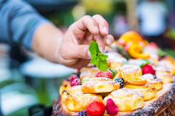 Chef prepares a plate of cakes with fresh fruits. He is working on the herb decoration. Outdoor garden party.