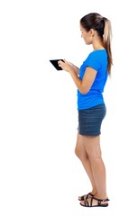 Naklejka premium back view of standing young beautiful woman using a mobile phone or tablet computer. girl watching. girl in a short skirt and a blue shirt stands sideways and manages a finger on the tablet.