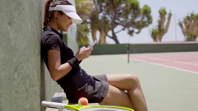 Attractive young female tennis player sitting on a bench at the side of an outdoor tennis court checking her mobile for text messages  side view
