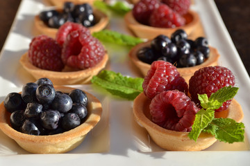 Mini fruit tarts with fresh raspberry and bilberry decorated mint leaves.. Delicious summer dessert in a porcelain plate. Rows of sweet treats. Party food.