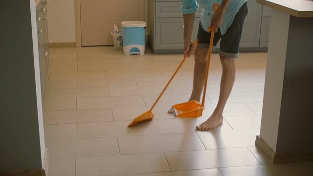 Young man, husband is goofing around with yellow broomstick while sweeping floor in the kitchen. Slow mo, Steadicam shot