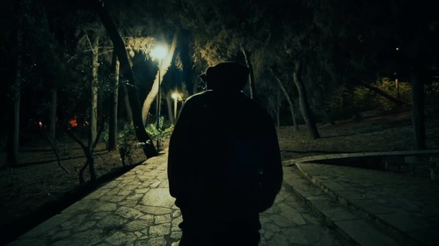 Suspicious hooded figure walks in a dark park at night