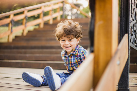 Portrait of little cute child sitting on wood stairs in city park. Model released cheerful caucasian boy looks at camera through wooden parapet of stairs. Horizontal color image.