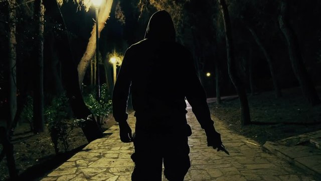 Hooded man walks in dark park draws knide,slomo 100p