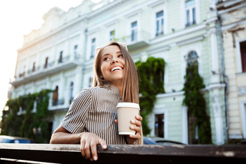 Young beautiful girl sitting on bench, holding coffee, smiling.