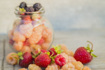Multicolored raspberries in a glass jar with strawberries on background