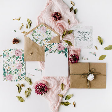 Workspace. Wedding invitation cards, craft envelopes, pink and red roses and green leaves on white background. Overhead view. Flat lay, top view