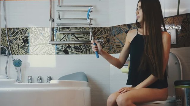 Girl sitting on toilet in bathroom. Take selfie on blue monopod. Posing. Apartment