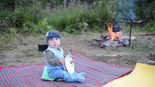 Cute kid sitting near a campfire and playing with plate and spoon