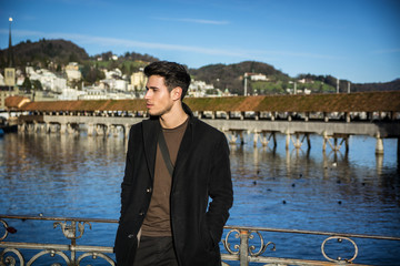 Fototapeta premium Handsome man standing near metal fence in Lausanne. Famous wooden bridge and Water Tower on background.