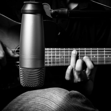 male musician playing acoustic guitar behind condenser microphone in recording studio, bw filter