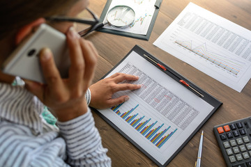 Business woman at workplace at wooden office table analyzes the data, schedules, prices, makes calculations on a calculator, talking on mobile phone