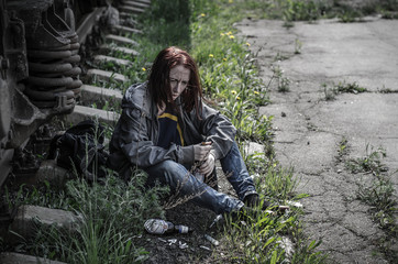 young woman in old dirty, ragged clothes sittingon the ground next to the wagon train and holding an empty bottle of alcohol, cigarettes and scattered around the used syringes