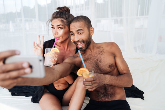 Multiracial couple making selfie and holding cocktails at the beach