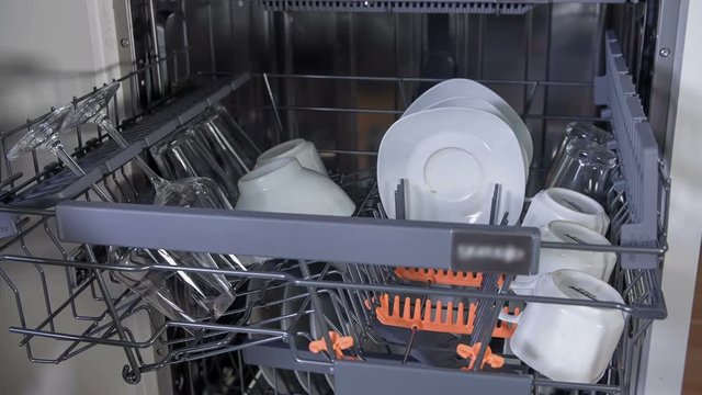 Woman is slowly placing saucers in the dishwasher, on the top rack and she is then putting the rack inside. Wide-angle shot.
