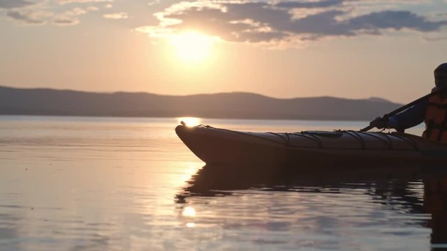 Side view of man slowly rowing a kayak with paddle on the lake in the evening at sunset