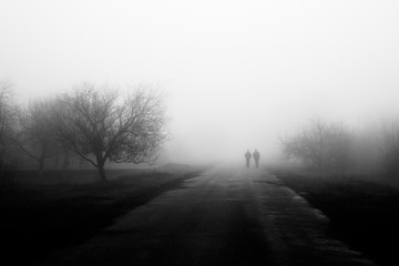 Wayfarers in fog. Silhouette of friends walking on misty village road. Homecoming. Loneliness,...