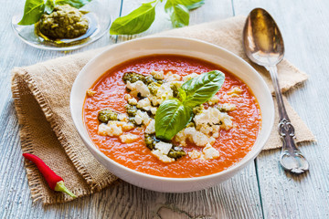 Tomato soup with feta cheese and pesto sauce in white bowl on wooden background. Fresh basil leaves and vegetables