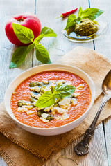 Tomato cream soup with feta cheese and pesto sauce in white bowl on wooden background. Fresh basil leaves and vegetables