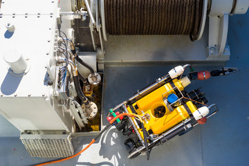 Small yellow mini submarine with gripping claw and rope attached seen from above. Tugging winch in background with winch machinery to the left.