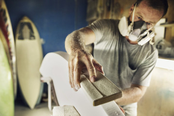 Mid adult man shaping edge of surfboard at workshop