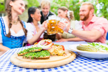 Fünf Freunde, Männer und Frauen, stoßen im Biergarten mit Bier an, bayerische Brotzeit steht auf...