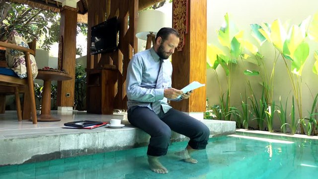Young businessman with documents sitting by pool in outdoor villa
