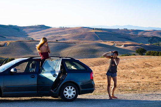 deux jeunes filles, sur le bord de la route et dans une voiture font des photos devant un paysage de Toscane en Italie