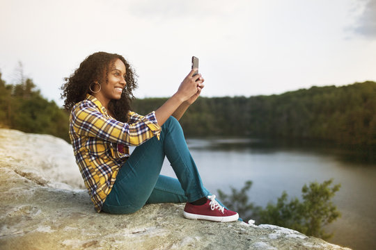 Happy woman taking selfie through smart phone on rock by lake