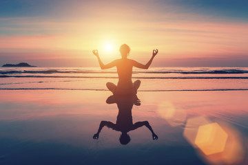 Female silhouette sitting in yoga pose on the beach by sea at sunset.