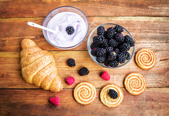 Products served for breakfast or snack, yoghurt in a glass bowl, croissant, cookies with sesame seeds and fresh ripe blackberry and raspberry on wooden surface, topview