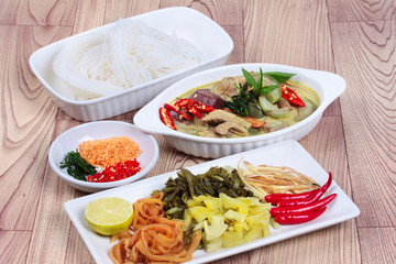 Rice noodle and green chicken curry in coconut milk with side dish on wood.