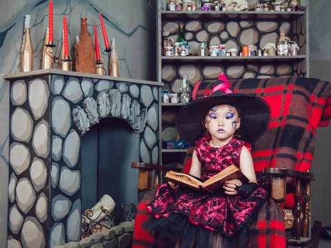 Girl in witch costume and make-up on her face sitting in a chair with  book in his hands in halloween decorations